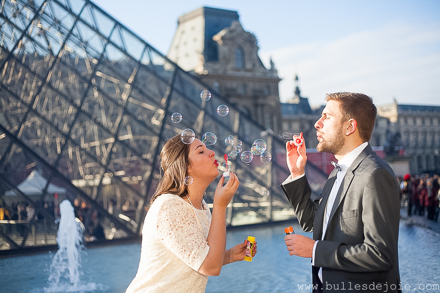 Couple jouant aux bulles de savon devant la Pyramides du Louvre | Séance photo romantique et fun | Bulles de Joie Photographie, photographe Paris